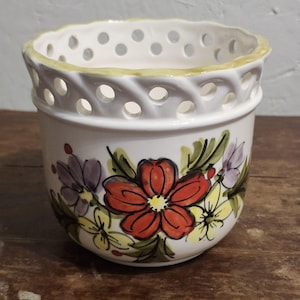 May include: A white ceramic planter featuring a hand-painted floral design with red, yellow, and purple flowers and green leaves. The upper rim has a decorative lattice cutout pattern with a scalloped edge finished in a light green paint.