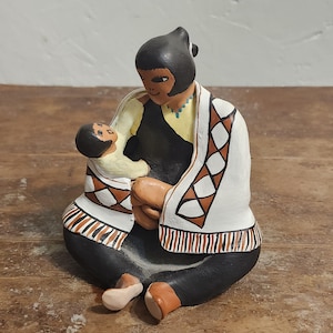May include: A ceramic figurine depicting a seated figure holding a baby. The figure wears a black dress, a white shawl with geometric patterns, and a black head covering. The baby is wrapped in a cream-colored blanket. The figurine is painted with earth-tone colors.