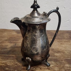 May include: A silver creamer with an ornate design and three feet. The creamer has a lid with a black knob.