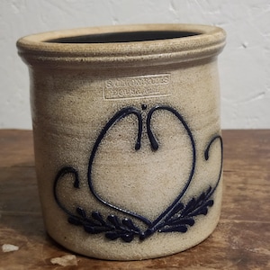 May include: A beige stoneware crock with a dark blue hand-painted heart and leaf motif. The vessel features a stamped rectangular mark reading SALMON FALLS DOVER, NH near the rim. The cylindrical container has a thick, rounded top edge.