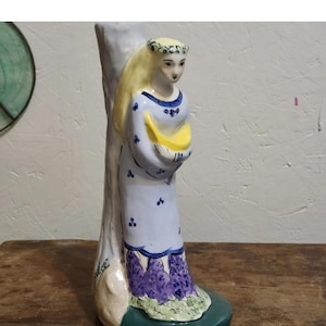 May include: A ceramic figurine of a person with long blonde hair and a floral crown, wearing a light blue dress with small blue floral patterns and a purple hem. The figure stands against a tall, light-colored tree trunk on a dark green base.
