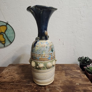 May include: A handcrafted ceramic vase with a flared, dark blue top and a textured, cylindrical body. The body features a painted landscape design, seashells, and turtle accents. The vase is approximately 12 inches tall.