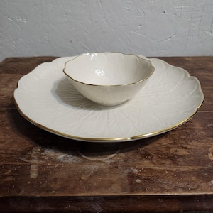 May include: A white ceramic serving plate and bowl set. The plate has a scalloped edge and a leaf pattern. The bowl sits in the center of the plate. Both pieces have a gold trim. The set is on a wooden surface.