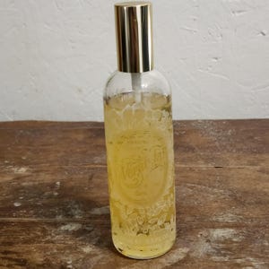 May include: A clear glass bottle with a gold-colored cap and a spray nozzle. The bottle is filled with a yellow liquid and has a decorative label with the text "Veranda Spa". The bottle contains 3.5 fl oz of liquid.