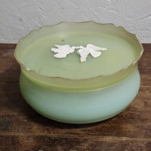 May include: A round, pale green glass bowl with a scalloped rim and a matching lid. The lid features two white, raised, molded bird figures facing each other in the center. The glass has a smooth, semi-translucent finish throughout the piece.