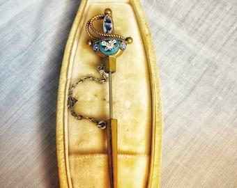 May include: A gold-toned sword-shaped pin with blue and white enamel detailing, in a velvet-lined box.