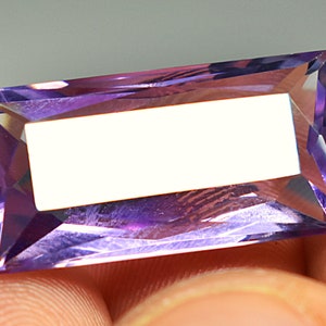May include: A rectangular, faceted amethyst gemstone with a purple hue. The gemstone is cut into a rectangular shape with sharp edges and a smooth, polished surface.
