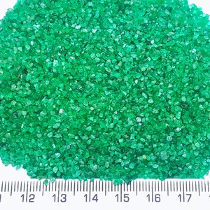 以下が含まれることがあります： 小さな緑色のエメラルドの宝石の山をクローズアップした写真。宝石は白い表面に広げられ、スケール用の定規が置かれています。定規はインチで測定値を示しています。