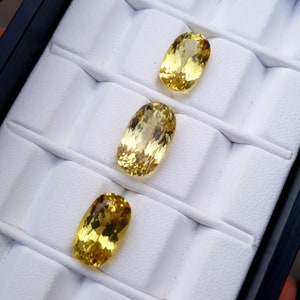 May include: Three oval-shaped yellow gemstones displayed in a white velvet-lined jewelry box.