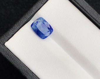 Puede incluir: Una vibrante gema azul con talla cojín rectangular, sobre fondo blanco en una caja negra. Las facetas de la gema capturan la luz, mostrando su claridad y color. Ideal para la creación de joyas.