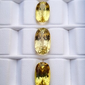 May include: Three oval-shaped yellow gemstones displayed in a white foam tray. The gemstones are different shades of yellow, ranging from pale to deep.