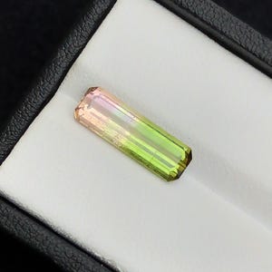 以下が含まれることがあります： 長方形のマルチカラーのトルマリン宝石。宝石は、片方の端のピーチ色から、ピンクがかった紫、鮮やかな緑へと変化します。石は、黒い箱の中の白い背景に置かれています。