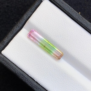 May include: A rectangular, multi-colored tourmaline gemstone. The gemstone transitions from pink to green, with a clear, polished appearance. It is displayed in a black jewelry box with a white, textured interior.