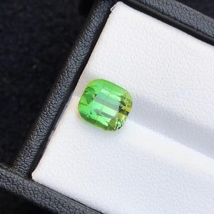 May include: A vibrant, square-cut, green gemstone displayed in a black and white jewelry box. The gemstone has a bright, translucent appearance, with visible internal facets. The box provides a contrasting background, highlighting the gem's color and clarity.