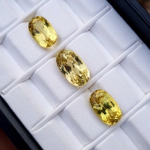 May include: Three oval-shaped yellow gemstones displayed in a white velvet jewelry box.