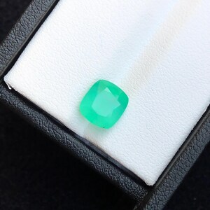 Puede incluir: Una vibrante gema verde esmeralda, de corte cuadrado, descansa en una caja blanca texturizada. La gema tiene una superficie facetada, que capta la luz. La caja es negra con un exterior texturizado, proporcionando contraste.