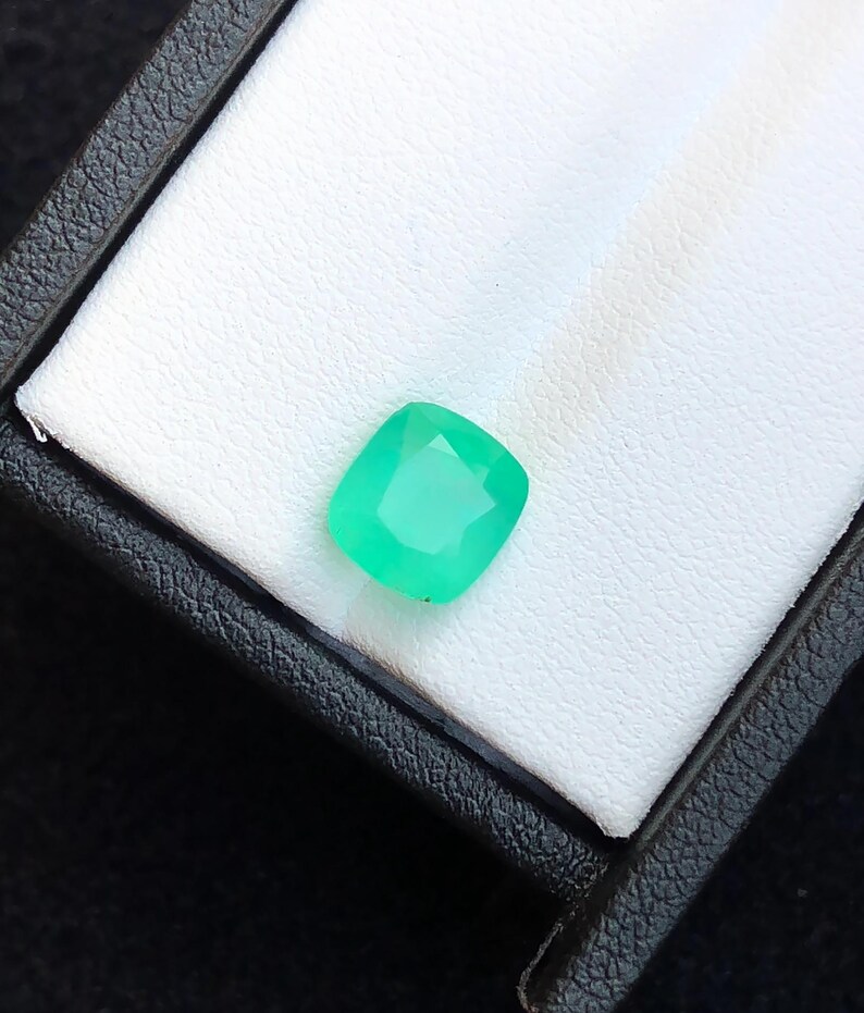 Puede incluir: Una vibrante gema verde esmeralda, de corte cuadrado, descansa en una caja blanca texturizada. La gema tiene una superficie facetada, que capta la luz. La caja es negra con un exterior texturizado, proporcionando contraste.