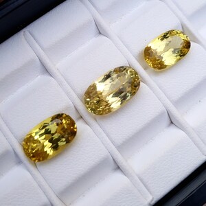 May include: Three oval-shaped yellow gemstones displayed in a white velvet jewelry box.