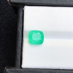 Puede incluir: Una esmeralda de talla coj&iacute;n, de color verde brillante, exhibida en una caja de joyer&iacute;a negra. La gema, con sus facetas visibles, se destaca sobre un fondo blanco. La caja proporciona un contraste.