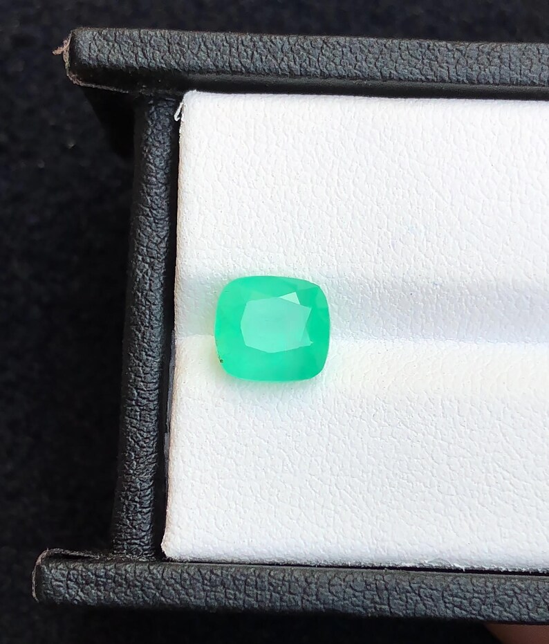 Puede incluir: Una esmeralda de talla coj&iacute;n, de color verde brillante, exhibida en una caja de joyer&iacute;a negra. La gema, con sus facetas visibles, se destaca sobre un fondo blanco. La caja proporciona un contraste.