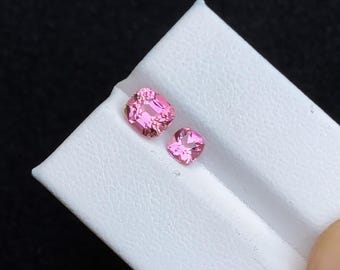 Puede incluir: Dos piedras preciosas cuadradas de color rosa sobre una superficie de exhibición blanca.
