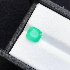 Puede incluir: Una vibrante esmeralda de talla coj&iacute;n, presentada en una caja de joyer&iacute;a negra con interior blanco. La gema es de un verde brillante y transl&uacute;cido, mostrando sus facetas. El marco proporciona un contraste, resaltando el color y la claridad de la gema.