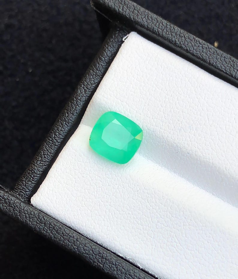 Puede incluir: Una vibrante esmeralda de talla coj&iacute;n, presentada en una caja de joyer&iacute;a negra con interior blanco. La gema es de un verde brillante y transl&uacute;cido, mostrando sus facetas. El marco proporciona un contraste, resaltando el color y la claridad de la gema.