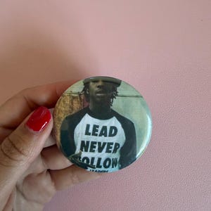 May include: A round button with a black and white image of a person wearing a baseball style shirt. The text "LEAD NEVER FOLLOW" is printed on the button. The word "LEADERS" is printed below the text.