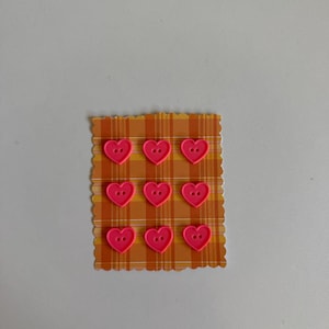 May include: Nine small, bright pink heart-shaped buttons with two holes in the center, arranged in a three-by-three grid on a rectangular piece of orange and yellow plaid paper with scalloped edges.