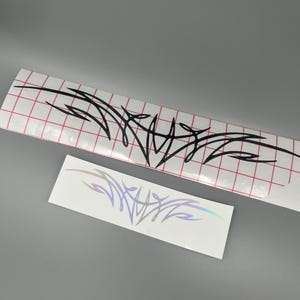 May include: Two adhesive decals featuring a sharp, abstract tribal-style design. The larger decal is black on a white backing with a red grid pattern, while the smaller decal is iridescent and reflective on a plain white backing.