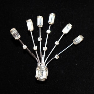 May include: A silver-toned brooch featuring a fan-like arrangement of six thin metal stems. Each stem is topped with a rectangular clear crystal, and smaller round clear crystals are set along the stems, anchored by a larger rectangular base crystal.