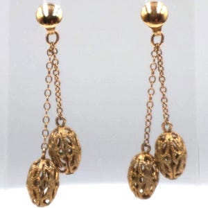 May include: A pair of gold-toned drop earrings featuring a smooth round stud connected to two delicate chains of varying lengths. Each chain terminates in an oval-shaped charm with an intricate, openwork filigree pattern throughout the surface.