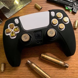 May include: A black PlayStation 5 controller with gold bullet-shaped buttons and triggers. The controller is surrounded by gold bullet casings on a wooden surface.