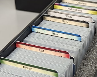May include: A black box with a foam insert containing a set of Magic: The Gathering cards organized by color. The colors are green, red, blue, black, white, multicolored, and colorless.