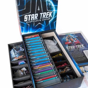 Puede incluir: Una caja de juego de mesa Star Trek Captain's Chair con un interior abierto que cuenta con divisores negros para organizar cartas, fichas y componentes. La tapa muestra el logo de Star Trek y un gráfico de un planeta azul, manual incluido.