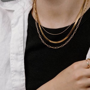 May include: A set of three gold chain necklaces layered on a person's neck. The chains are different widths and styles. The person is also wearing two gold bracelets on their wrist.