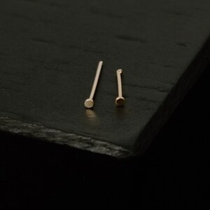 May include: Two gold nose rings on a black surface. The nose rings are thin and have a small, round bead at the end.