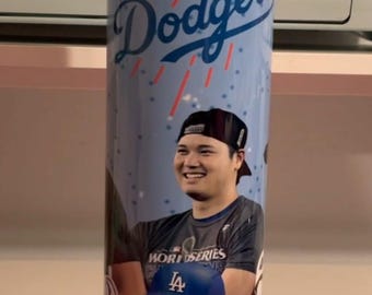 Op de afbeelding: Een blauwe en witte beker met een baseball-ontwerp en het woord "Dodgers" in blauw. De beker toont afbeeldingen van twee honkballers, een draagt een grijs shirt met de woorden "World Series" en de ander draagt een blauwe honkbalpet met de letter "LA" erop.
