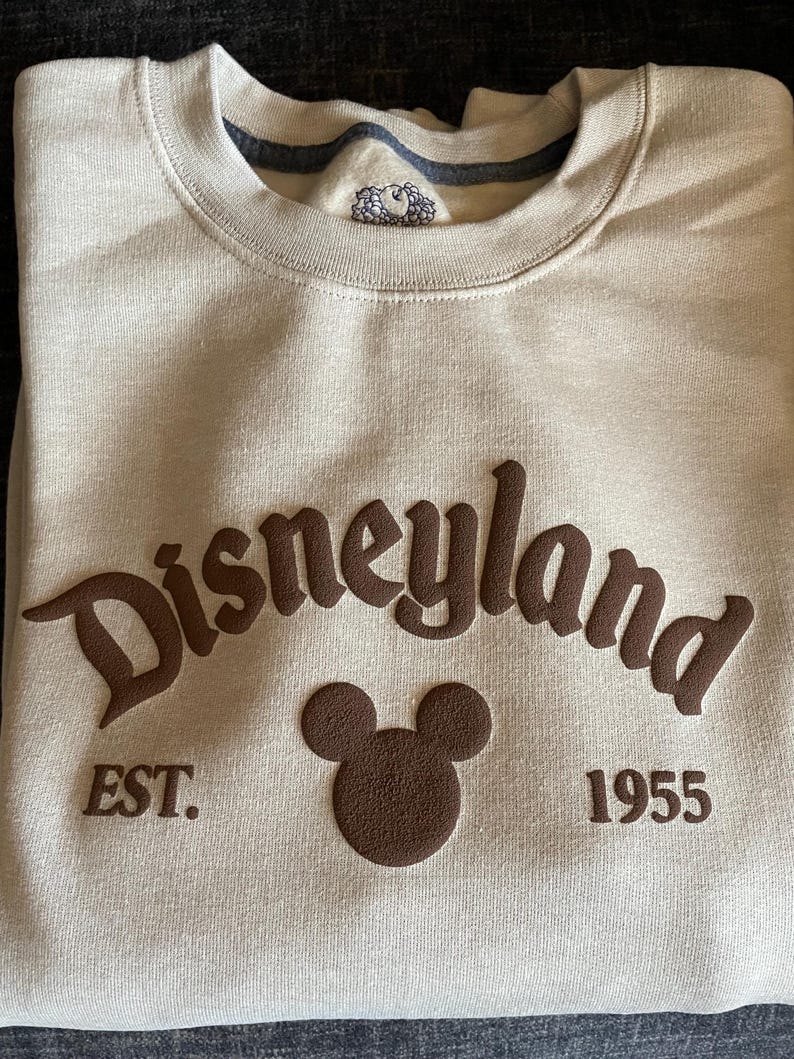 May include: A beige sweatshirt with the word "Disneyland" in brown, stylized lettering. Below the word "Disneyland" is a brown Mickey Mouse head and the text "EST. 1955". The sweatshirt is folded.