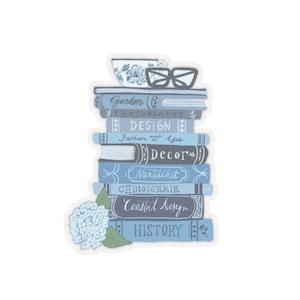 Puede incluir: Una pila de libros azules y blancos con títulos como "Garden", "Photography", "Design", "Fashion", "Decor", "Nantucket", "Chinoiserie", "Coastal Design" e "History". Una taza de té azul y blanca y un par de gafas están encima de la pila.