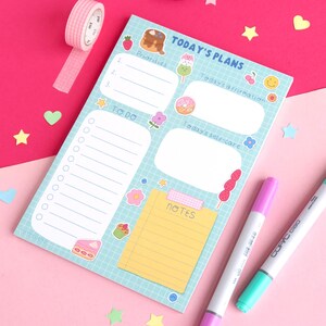 May include: A blue notepad with a grid pattern and colourful cartoon illustrations of food and flowers. The notepad has sections for "Today's Plans", "Priorities", "To Do", "Today's Affirmation", "Today's Self-Care", and "Notes".