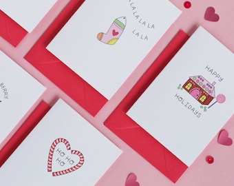 Puede incluir: Cinco tarjetas de Navidad con diferentes ilustraciones y saludos. Las tarjetas son blancas con contornos negros y acentos coloridos. Los saludos incluyen "Have a Berry Christmas", "Ho Ho Ho", "La La La La La", "Felices Fiestas", y una casa de jengibre con un bastón de caramelo.