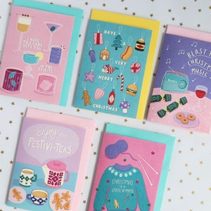 Pu&ograve; includere: Cinque biglietti d'auguri di Natale con diversi design. Le carte sono rosa, blu, gialle, viola e verdi. Le carte presentano illustrazioni di cocktail, ornamenti, tazze da t&egrave;, un maglione e una radio.