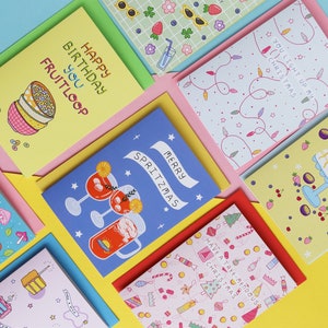 Pu&ograve; includere: Una collezione di biglietti d'auguri colorati con varie illustrazioni e testi. I biglietti presentano disegni come una ciotola di cereali, un albero di Natale, una torta e un bicchiere di spritz. Il testo sui biglietti include "Happy Birthday You Fruitloop", "You Light Up My Christmas", "I Pav-Love-Ya", "Merry Spritzmas", e "Have a Tree Mendous Christmas".