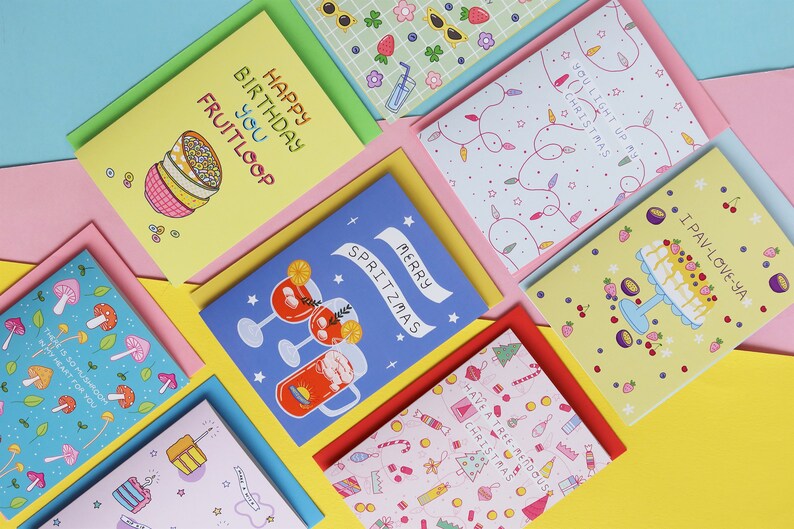 Pu&ograve; includere: Una collezione di biglietti d'auguri colorati con varie illustrazioni e testi. I biglietti presentano disegni come una ciotola di cereali, un albero di Natale, una torta e un bicchiere di spritz. Il testo sui biglietti include "Happy Birthday You Fruitloop", "You Light Up My Christmas", "I Pav-Love-Ya", "Merry Spritzmas", e "Have a Tree Mendous Christmas".