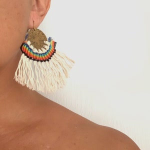 Puede incluir: Un par de pendientes con un dije redondo de latón y una franja de hilo blanco. El dije está decorado con una banda tejida de colores arcoíris.