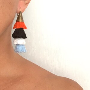 Puede incluir: Un par de pendientes de borla en capas con una parte superior de cono dorado. Las borlas son de color naranja, negro, blanco y azul claro.