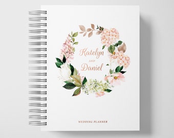 May include: A white wedding planner with a floral wreath design. The wreath is made of pink and white flowers, green leaves, and brown branches. The text "Katelyn and Daniel" is written in rose gold lettering. The words "Wedding Planner" are printed in black at the bottom of the cover.