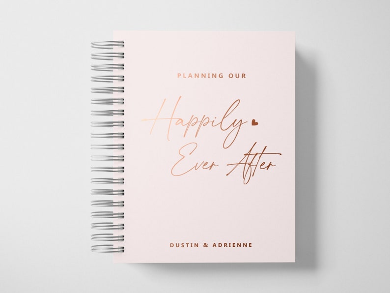 May include: A pink spiral bound notebook with the text "Planning Our Happily Ever After" in rose gold lettering. The names "Dustin & Adrienne" are printed in rose gold at the bottom of the cover.