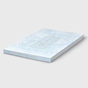 May include: A light blue baby shower guest book with a floral pattern. The book has gold lettering that reads "Emily's Baby Shower" and the date. The book is closed and lying on a white surface.